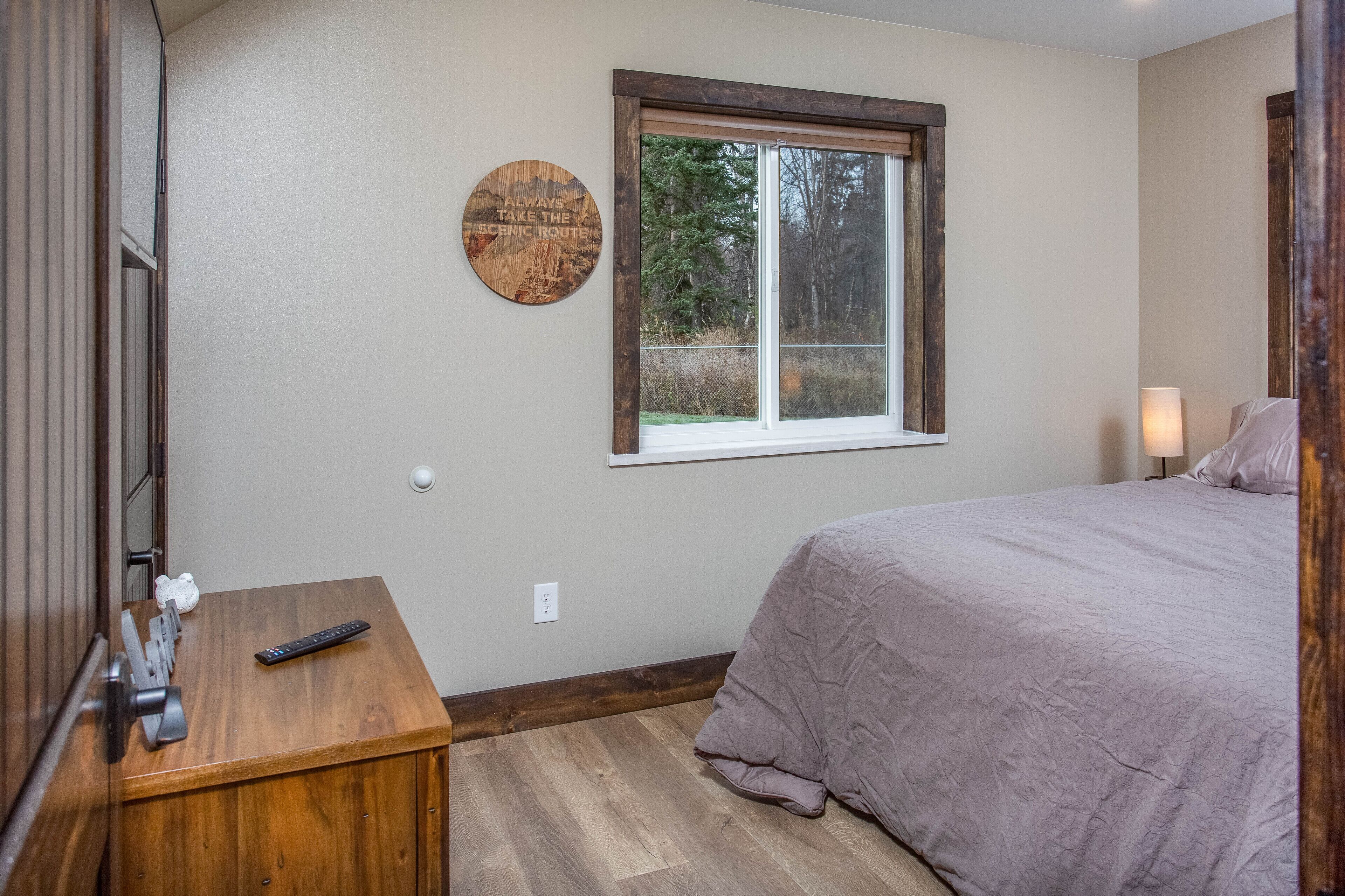 A bedroom with a bed, a wooden nightstand, a wall-mounted clock, and a window with a view of trees.