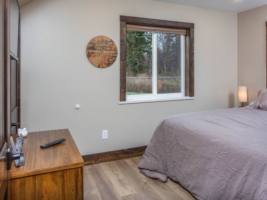 A bedroom with a bed, a wooden nightstand, a wall-mounted clock, and a window with a view of trees.