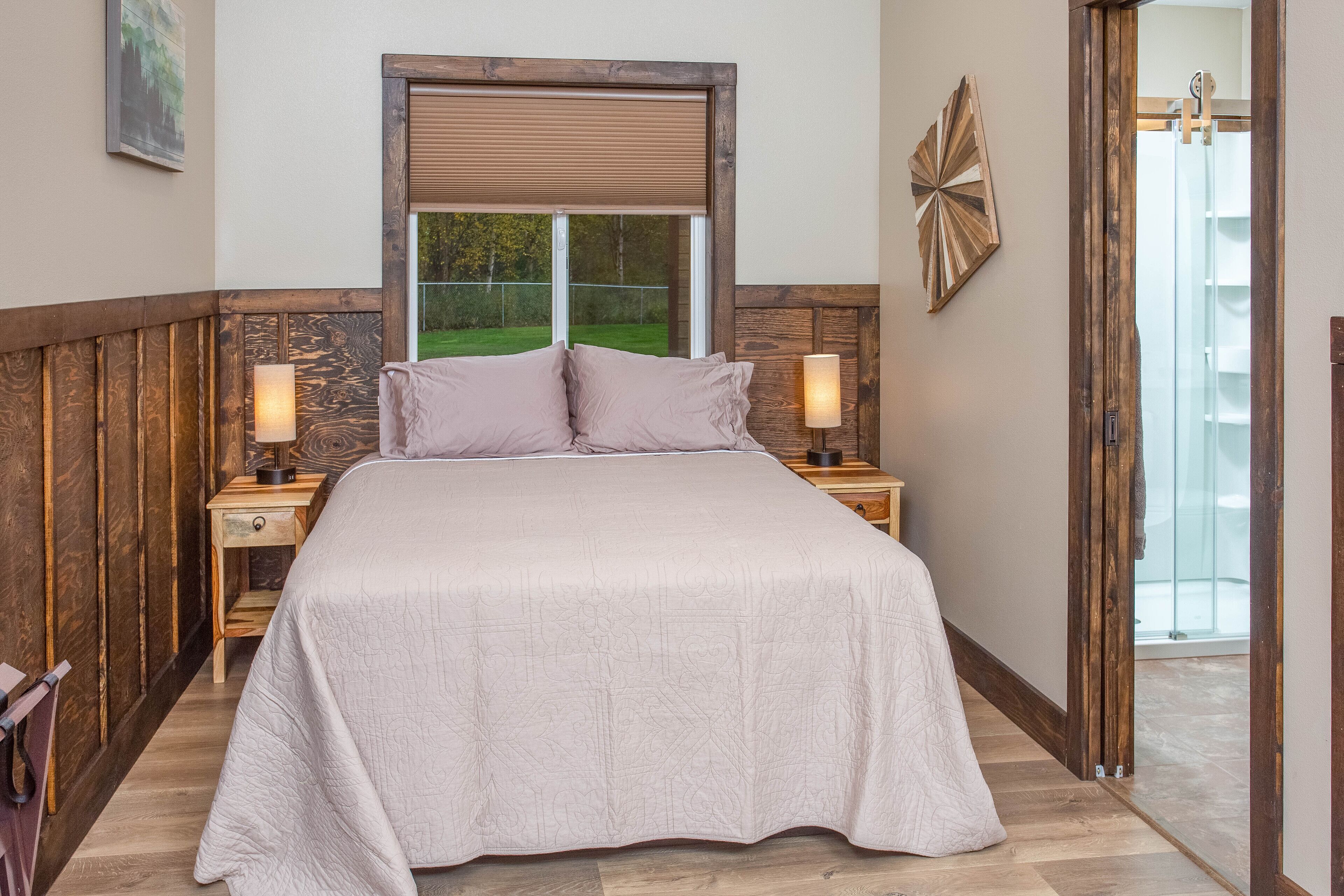 Quarto com cama grande, cabeceira de madeira, duas mesas de cabeceira com luminárias, janela com persianas e vista para uma paisagem verde.