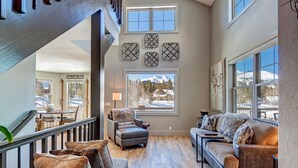 A spacious living area with a high ceiling, a staircase, and large windows offering a view of a snowy mountain. - Creekside Escape: Breck Home, Hot Tub & Steam Shower, Walk to Shuttle! (Breckenridge)
