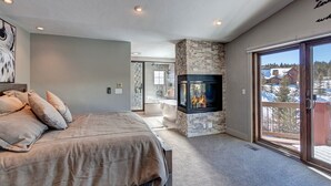 A bedroom with a large bed, a stone fireplace, a sliding glass door to a balcony, and a bathroom with a bathtub. - Creekside Escape: Breck Home, Hot Tub & Steam Shower, Walk to Shuttle! (Breckenridge)