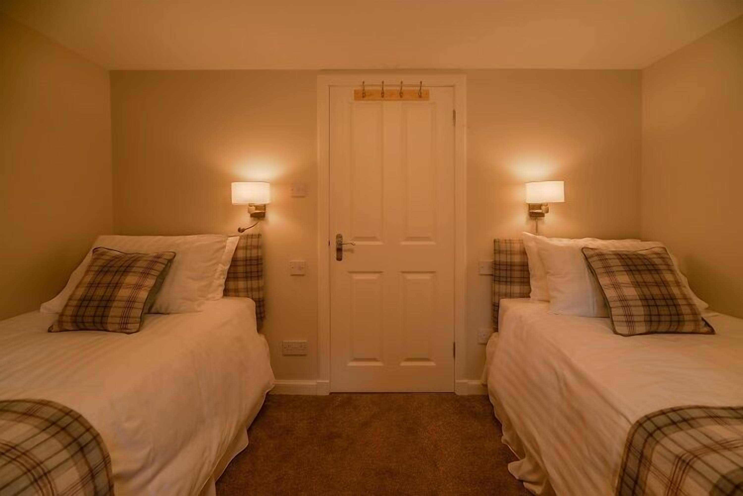 A room with two beds, each with a plaid pillow and a bedside lamp.