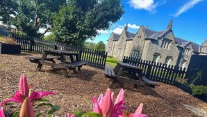 A stone building with a traditional architectural style, surrounded by a wooden fence and a picnic table area. - Blairquhan Cottages (Maybole)