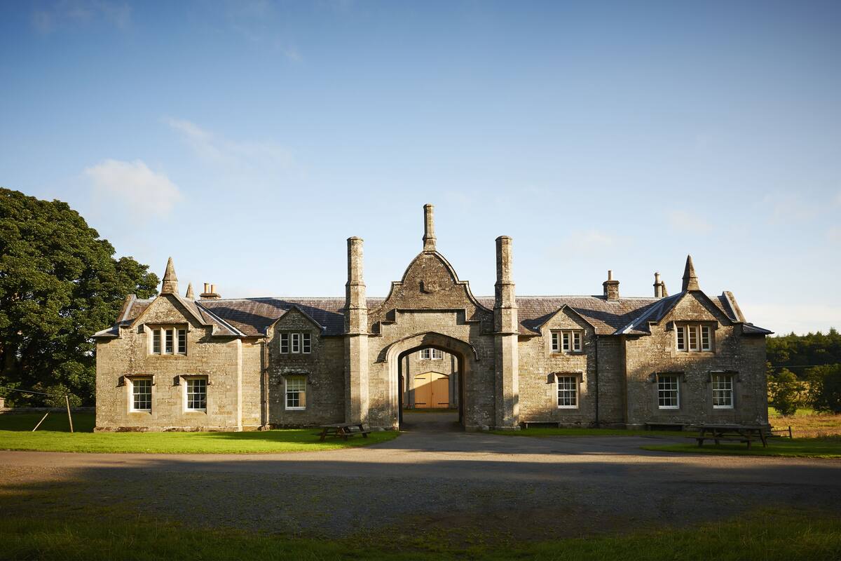 A large stone building with a central archway, multiple chimneys, and a paved driveway.