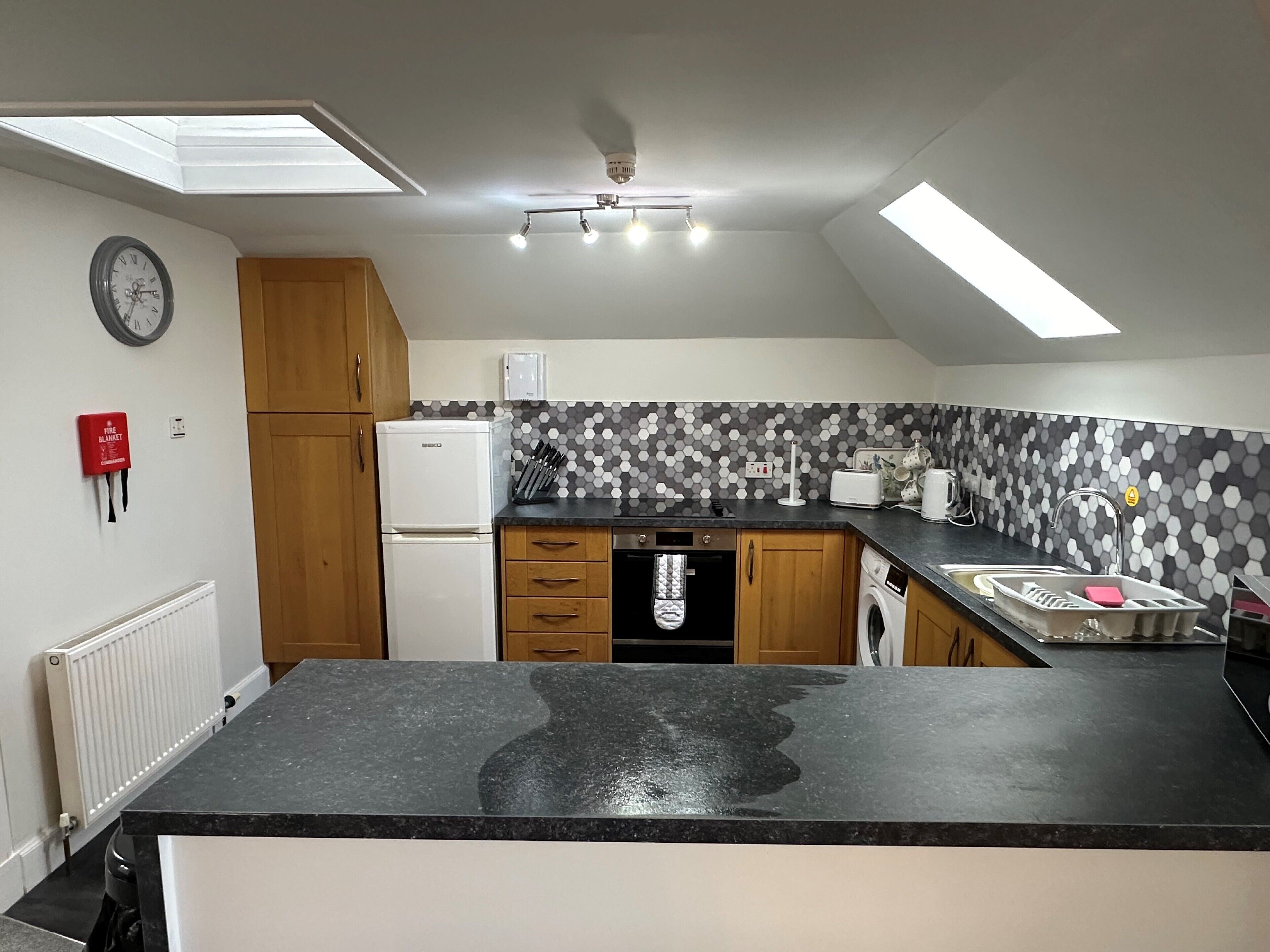 A kitchen with a black countertop, wooden cabinets, a white refrigerator, a microwave, and a tiled backsplash.
