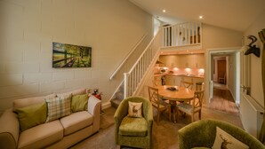 A cozy living room with a sofa, armchair, and dining table. A staircase leads to an upper level with a kitchen and a hallway. - Blairquhan Cottages (Maybole)