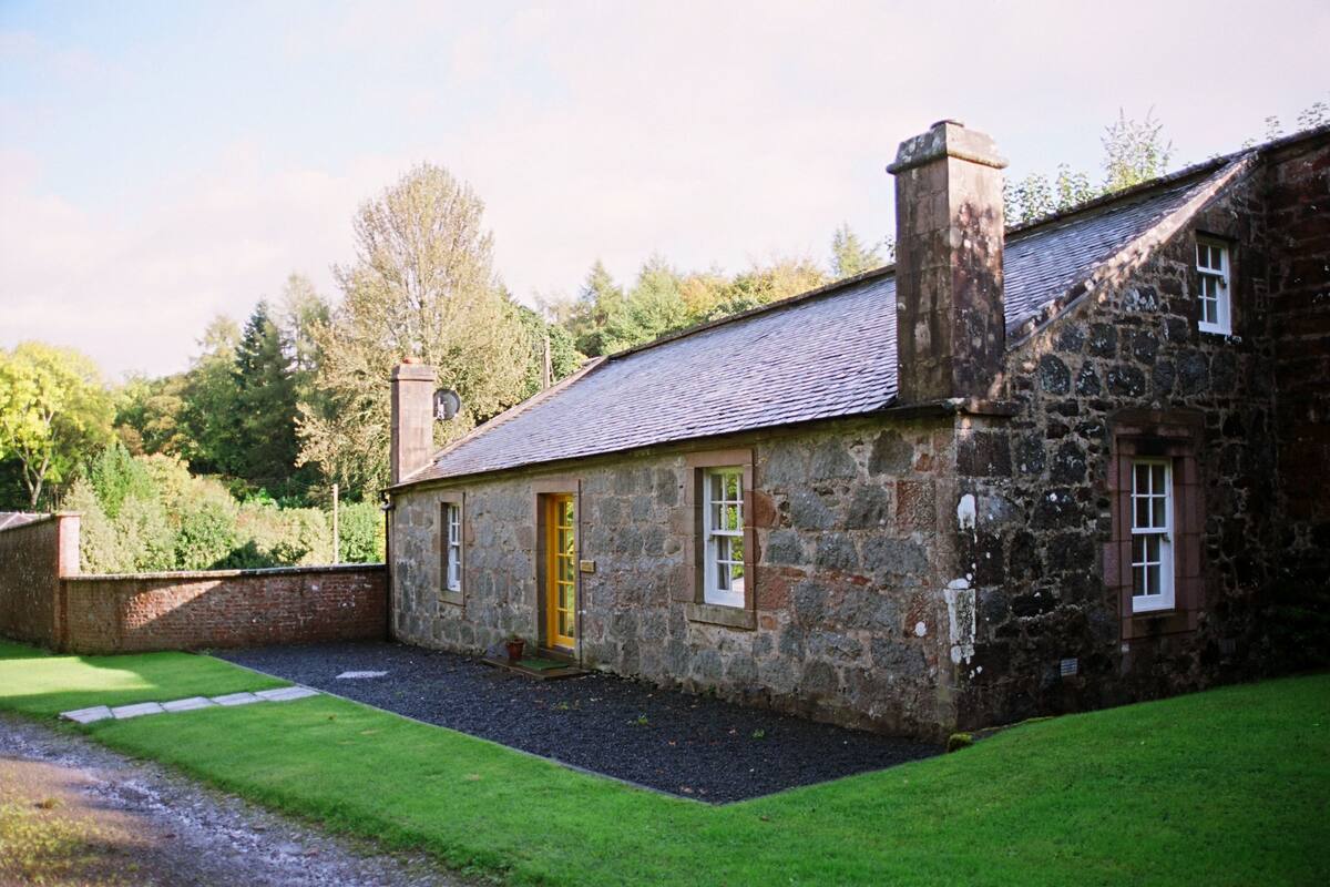 A stone house with a sloped roof, a chimney, and a small front yard with a pathway leading to the entrance.