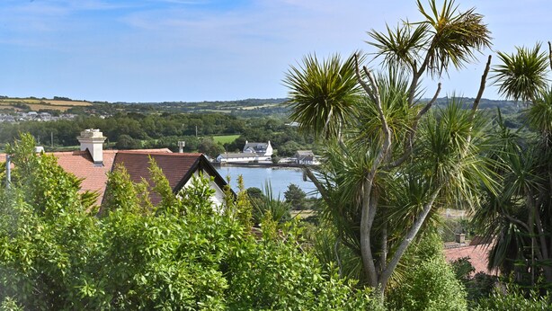 A coastal view with a building featuring a red-tiled roof, surrounded by lush greenery and palm trees. - Halwell Lodge (St Ives)