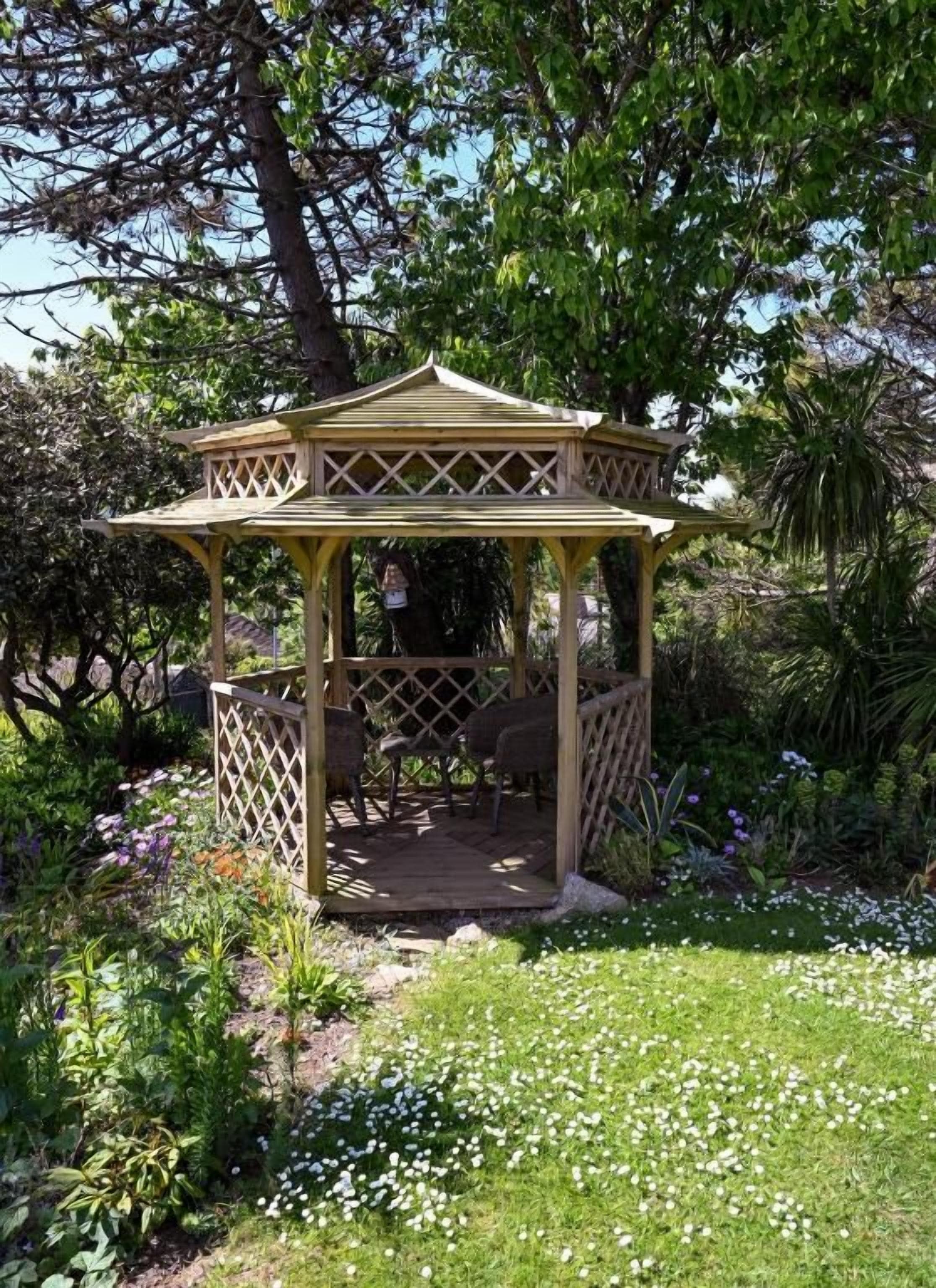 Un pabellón de madera con techo de celosía, rodeado de un jardín con diversas plantas y flores.