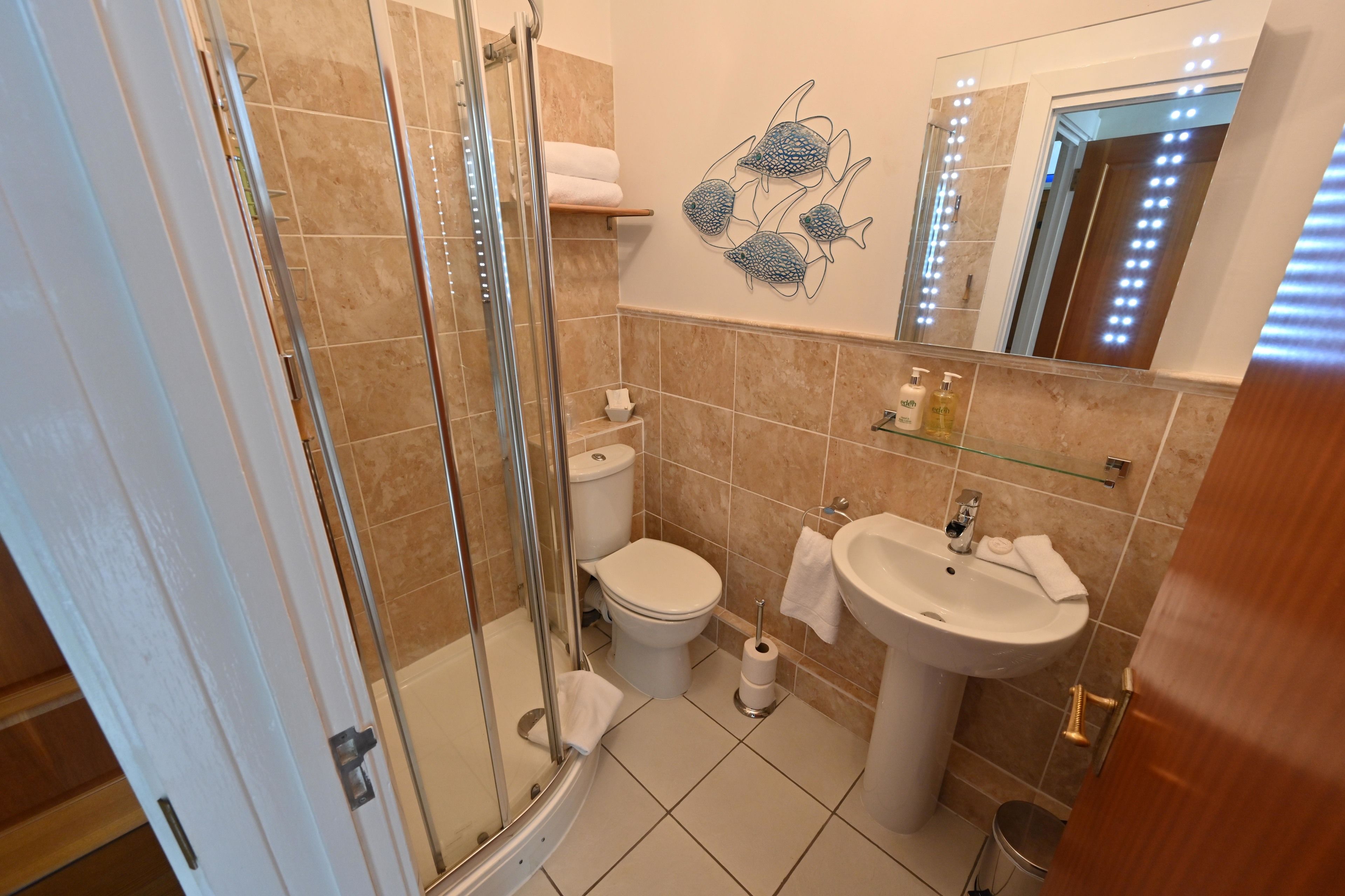 A bathroom with a glass shower enclosure, a toilet, a sink, and a mirror with lights.