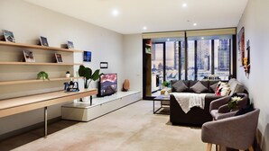 A modern living room with a city view, a sofa, a coffee table, and a TV on a stand. - Short Stay Apartment at Flinders (Melbourne)