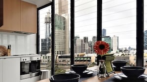 A modern kitchen with large windows, a dining table with black bowls and a vase of red flowers, and a view of a cityscape. - Short Stay Apartment at Flinders (Melbourne)