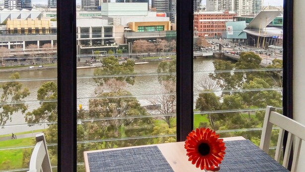 A cityscape view from a window with a table featuring a red flower. - Short Stay Apartment at Flinders (Melbourne)