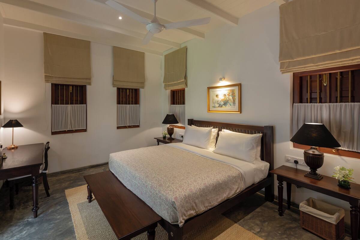 A bedroom with a bed, bedside tables, a bench, a ceiling fan, and a painting on the wall.