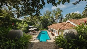 A swimming pool surrounded by lush greenery, with a tiled-roofed building and a large decorative vase in the foreground.