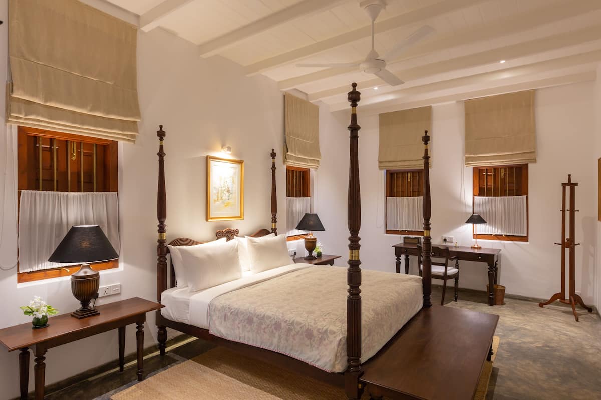 A four-poster bed with a wooden frame, a bedside table with a lamp, a desk with a chair, and a standing lamp.