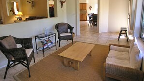 Een ruime woonkamer met een houten salontafel, twee rieten fauteuils, een bank en een eethoek op de achtergrond.