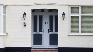 Une maison blanche avec une porte à deux battants, deux fenêtres avec volets et une plaque portant le numéro « 16 ».