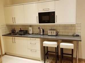 Eine kleine Küche mit einer Mikrowelle, einem Toaster, einem Wasserkocher und einem Spülbecken. Zwei Barhocker und ein Schrank mit Schubladen sind vorhanden.