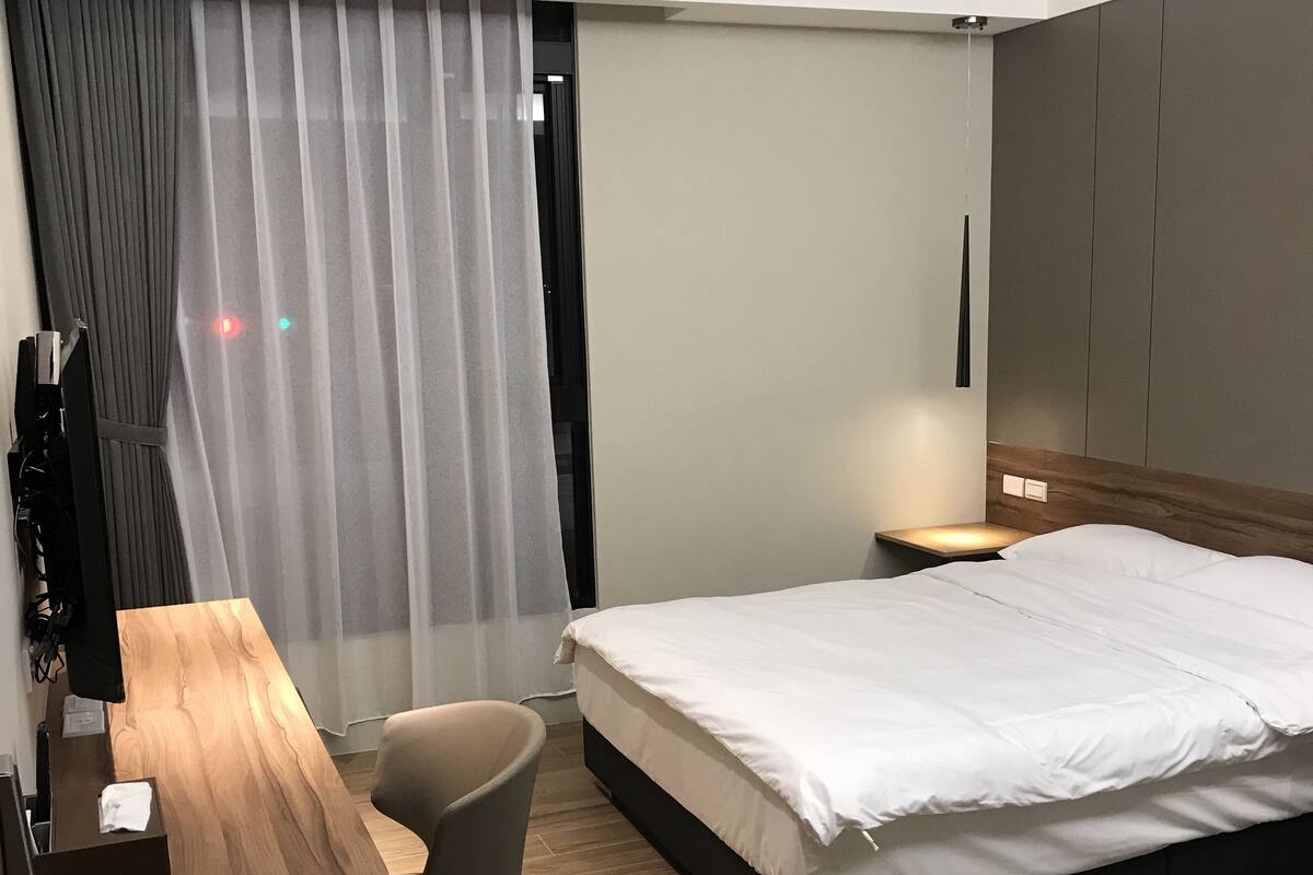 A hotel room with a bed, a desk, a chair, an air conditioning unit, a curtain, and a nightstand with a lamp.