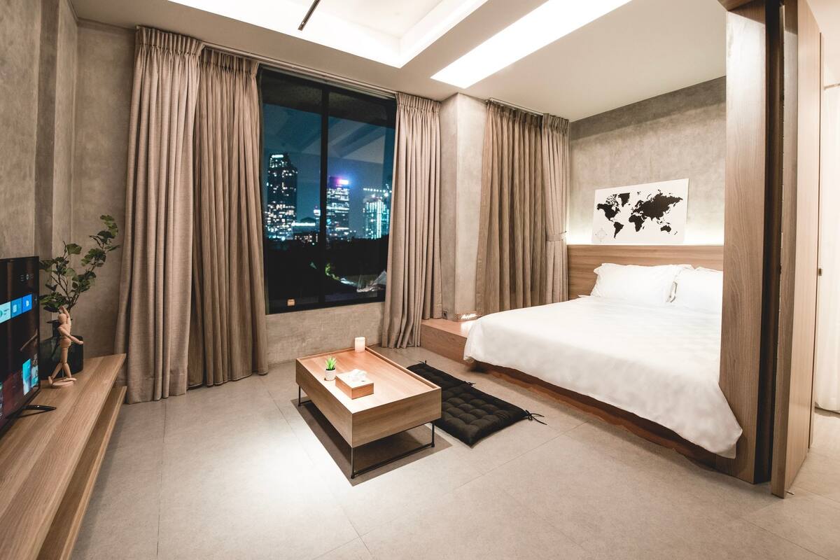 A modern hotel room with a large bed, a wooden coffee table, a TV, and a view of a cityscape at night.