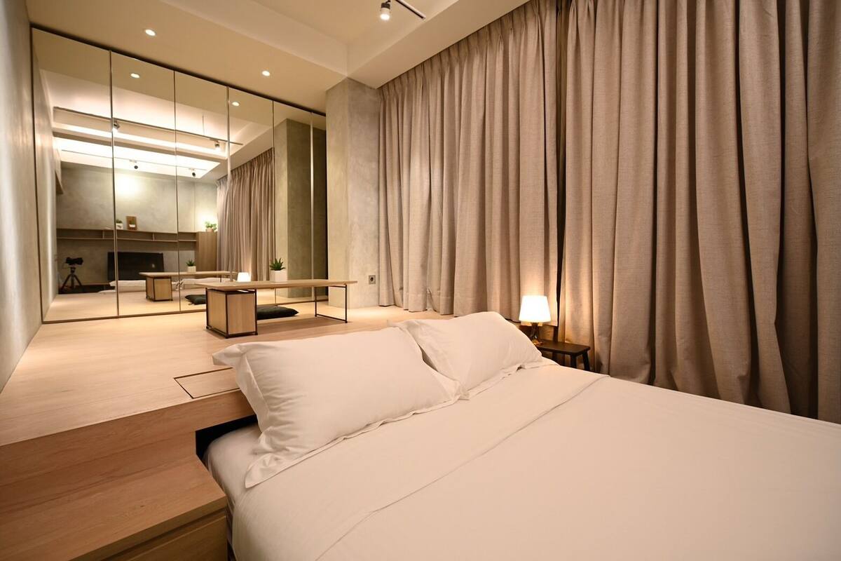 A modern hotel room with a large bed, a bedside table, a nightstand with a lamp, a mirror, and a view of a dining area with a table and chairs.