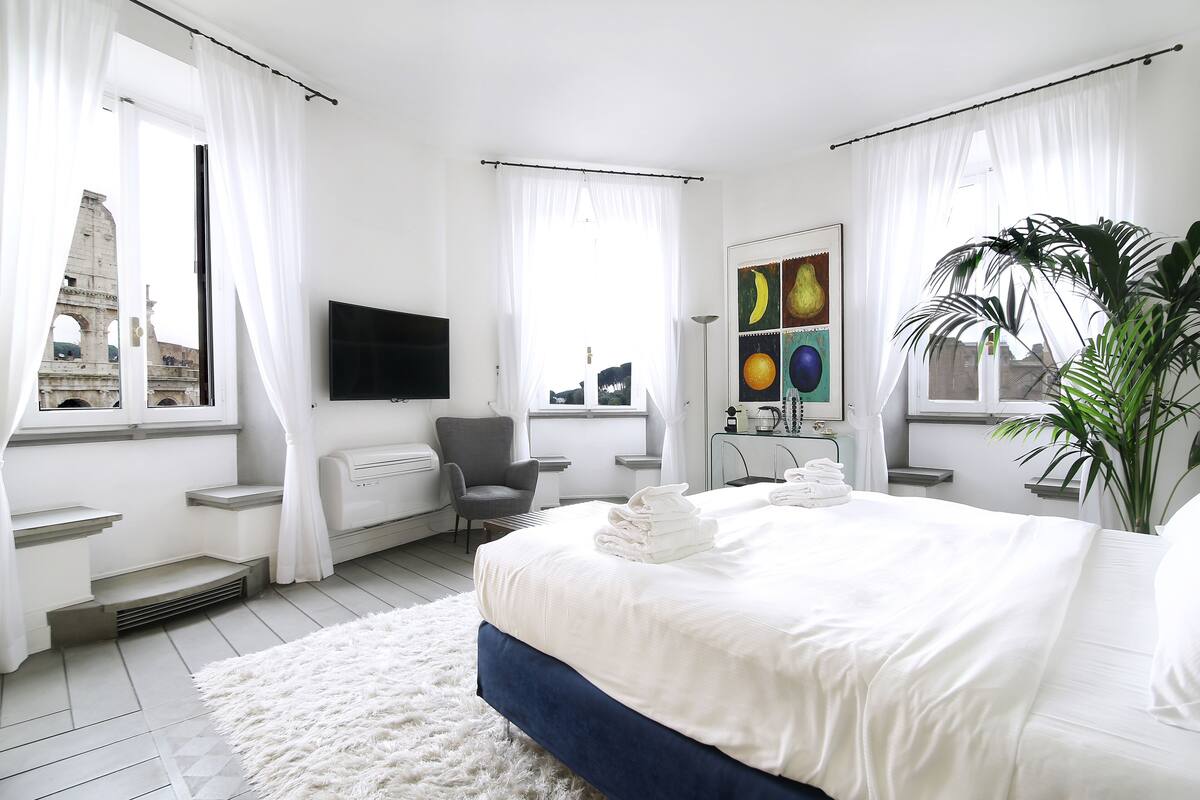 Quarto de hotel moderno com uma cama grande, televisão de tela plana, escrivaninha e vista para um edifício histórico pela janela.