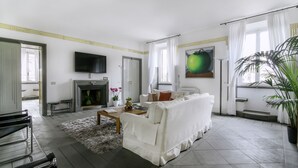 A modern living room with a white sofa, a flat-screen TV, a fireplace, and a large window with curtains. - Magia at Colosseum (Rome)