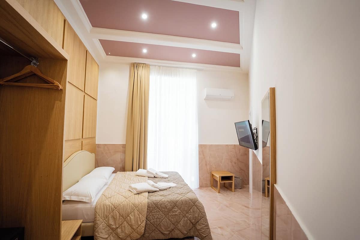 Une chambre d’hôtel avec un lit, une armoire en bois, un petit tabouret en bois, un téléviseur fixé au mur et une fenêtre avec des rideaux.