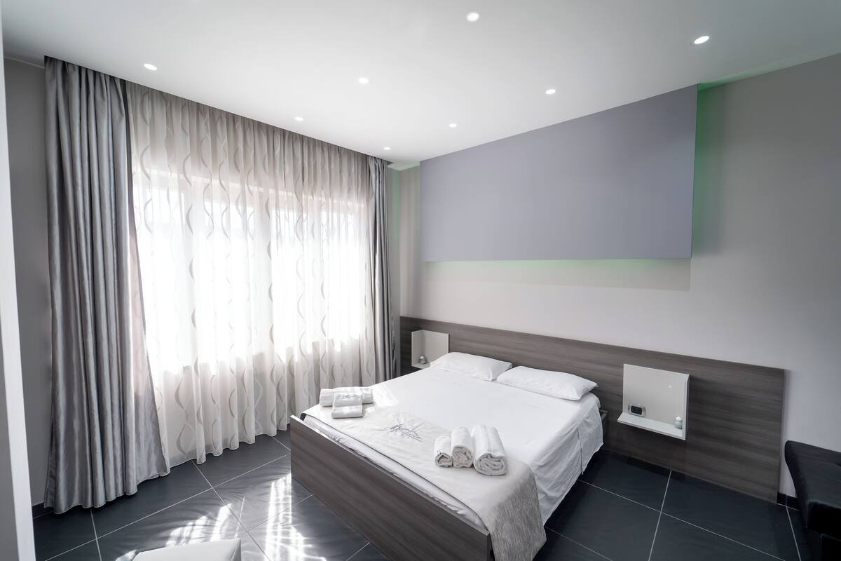Une chambre d’hôtel moderne dotée d’un grand lit, d’une table de chevet, d’un téléviseur fixé au mur et d’une fenêtre avec des rideaux légers.
