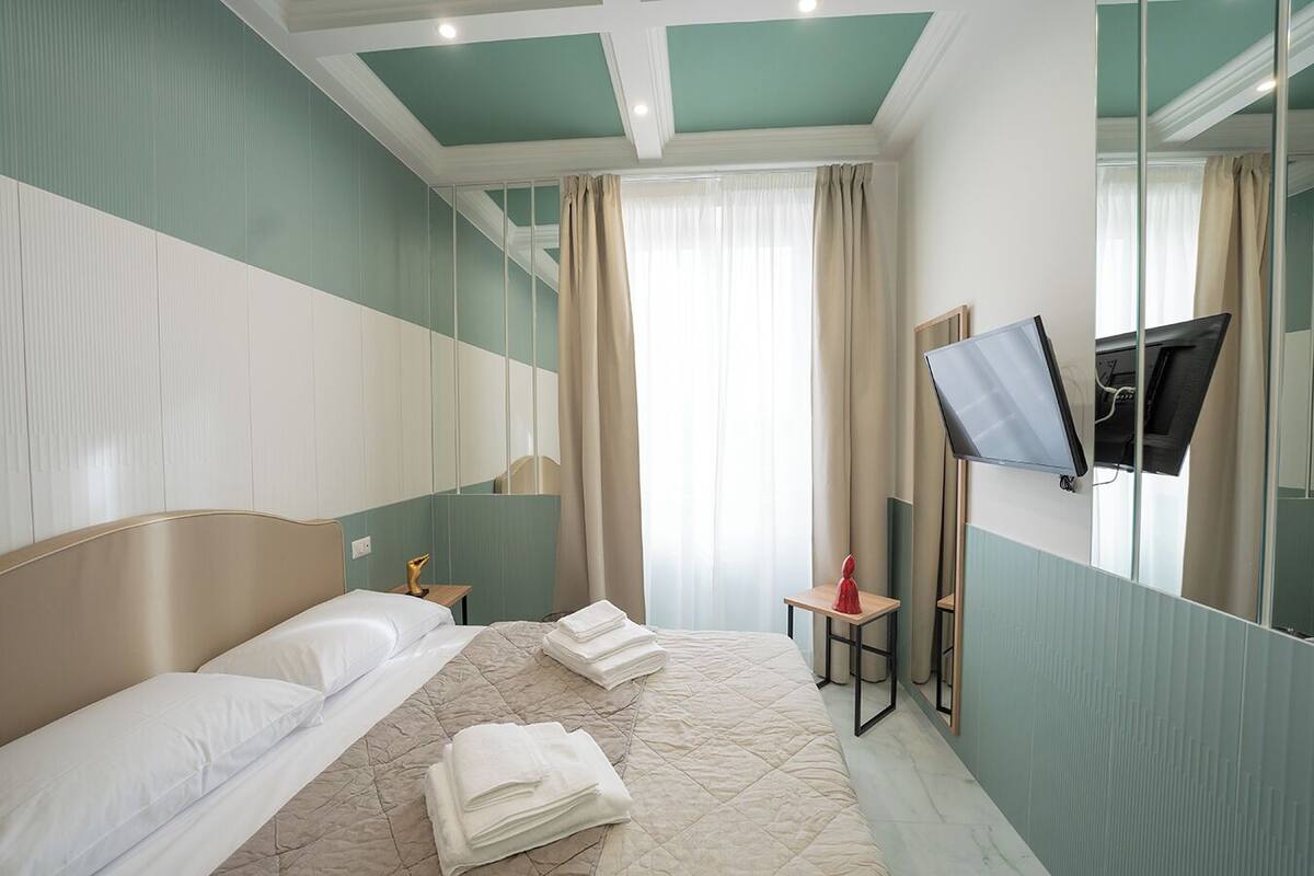 Une chambre d’hôtel avec un lit, un téléviseur fixé au mur, un miroir et une fenêtre avec des rideaux.
