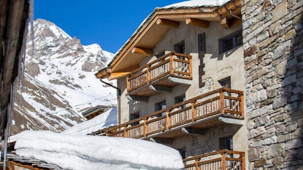 A multi-story building with wooden balconies and stone walls, set against a backdrop of snow-covered mountains. - Chalet Hôtel du Fornet (Val-d'Isere)