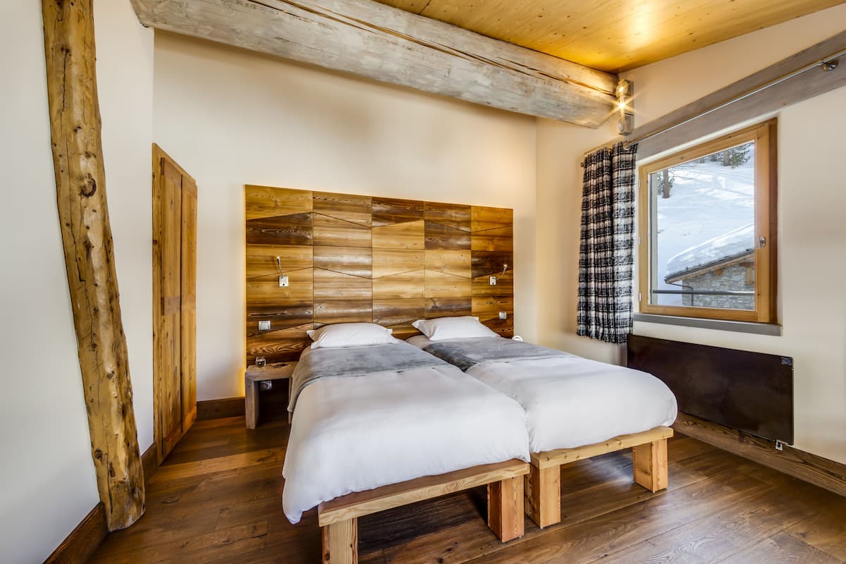 Una habitación con cabecera de madera, dos camas, un banco, una ventana con vista a la nieve y suelo de madera.