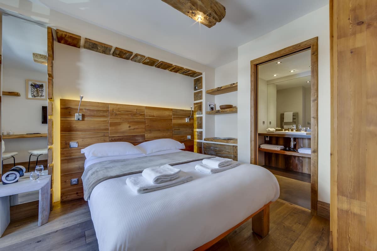 Un dormitorio moderno con cabecera de madera, cama bien hecha con toallas, mesita de noche con vasos y un libro, y un espejo en el baño contiguo.