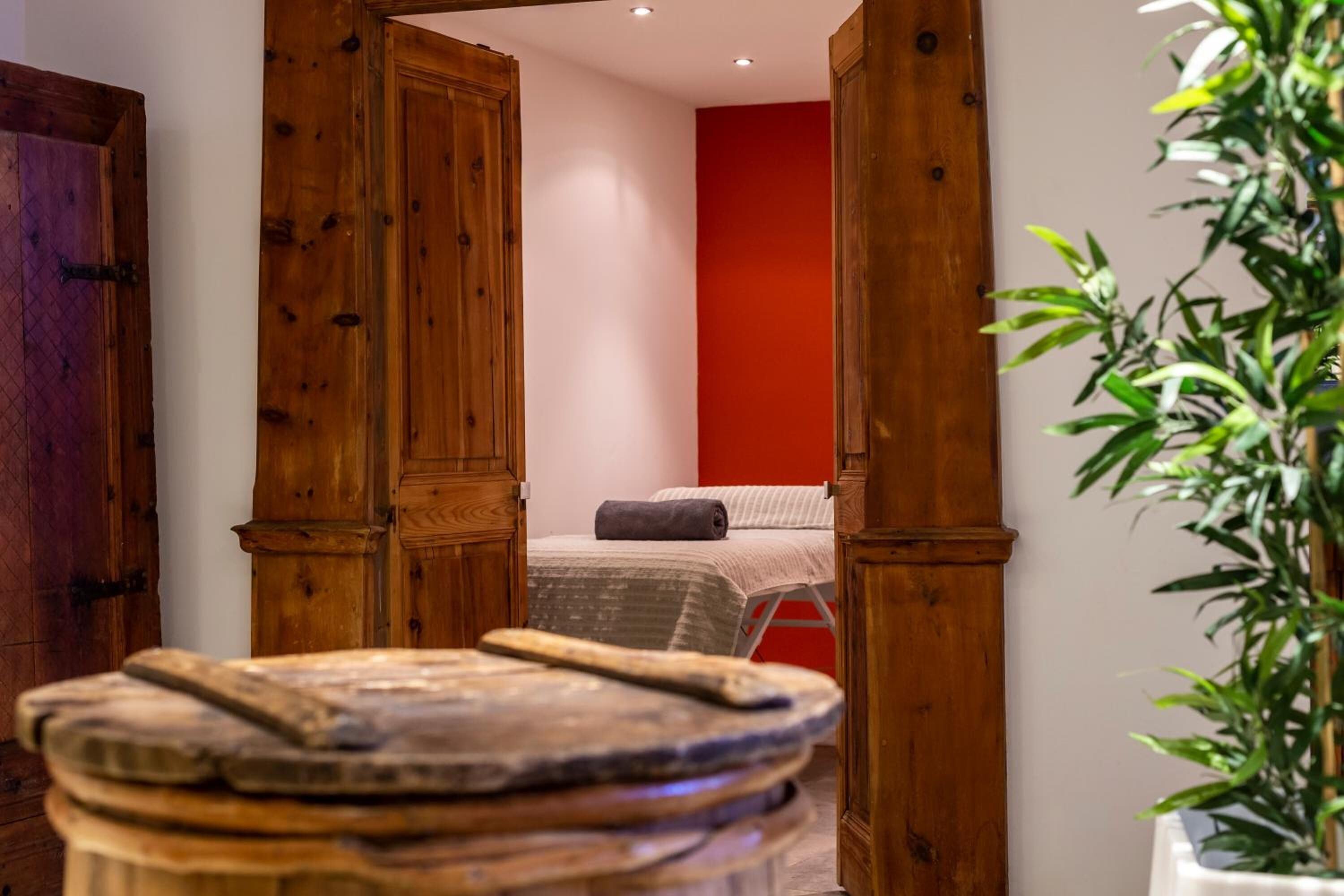 A narrow hallway with wooden doors leading to a room with a bed and a red wall. A plant is visible in the foreground.