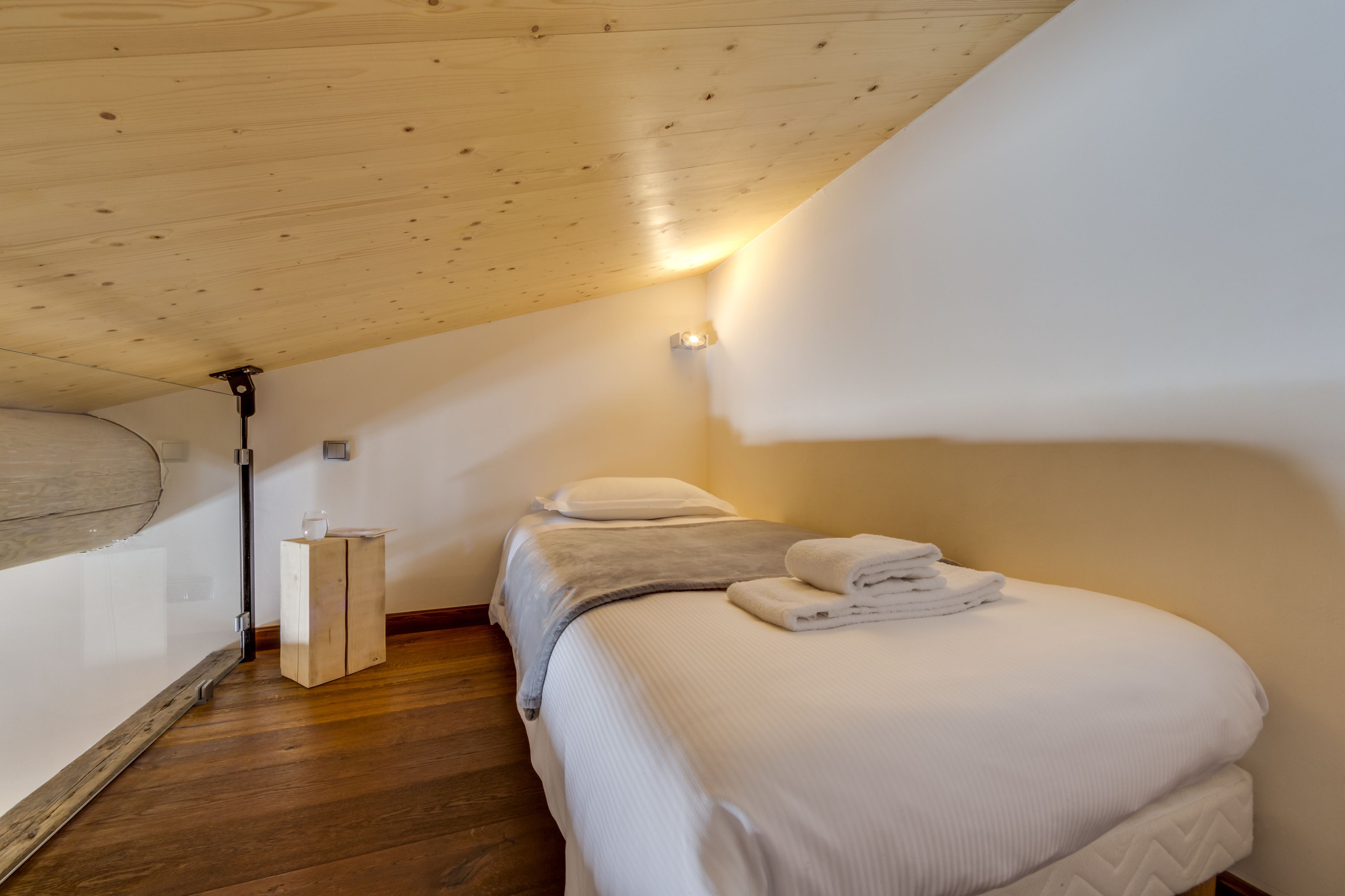 A bedroom with a sloped ceiling, wooden flooring, a bed with white bedding, and a small bedside table with a glass.