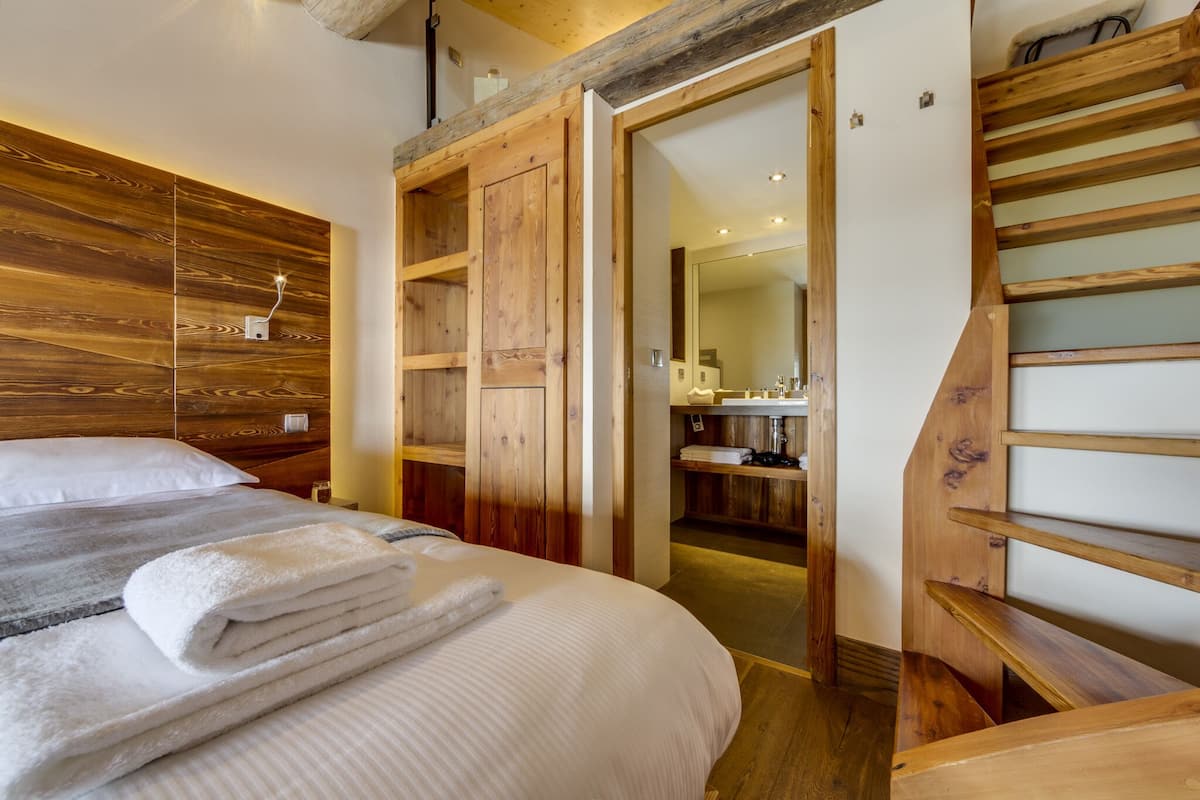 Habitación de hotel con cabecera de madera, cama con ropa blanca, una escalera de madera y un baño visible a través de una puerta abierta.