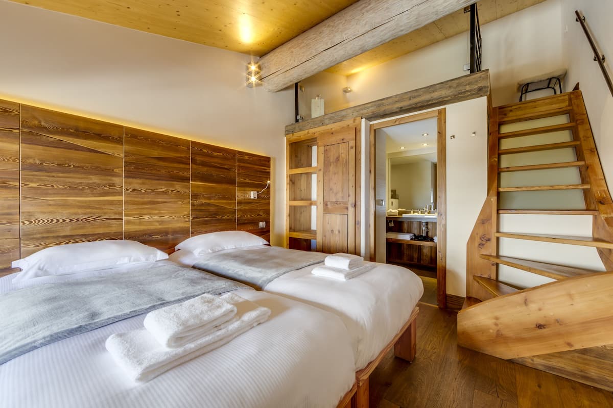 Habitación de hotel con dos camas, paneles de madera en las paredes, una escalera de madera y un baño visible a través de una puerta abierta.
