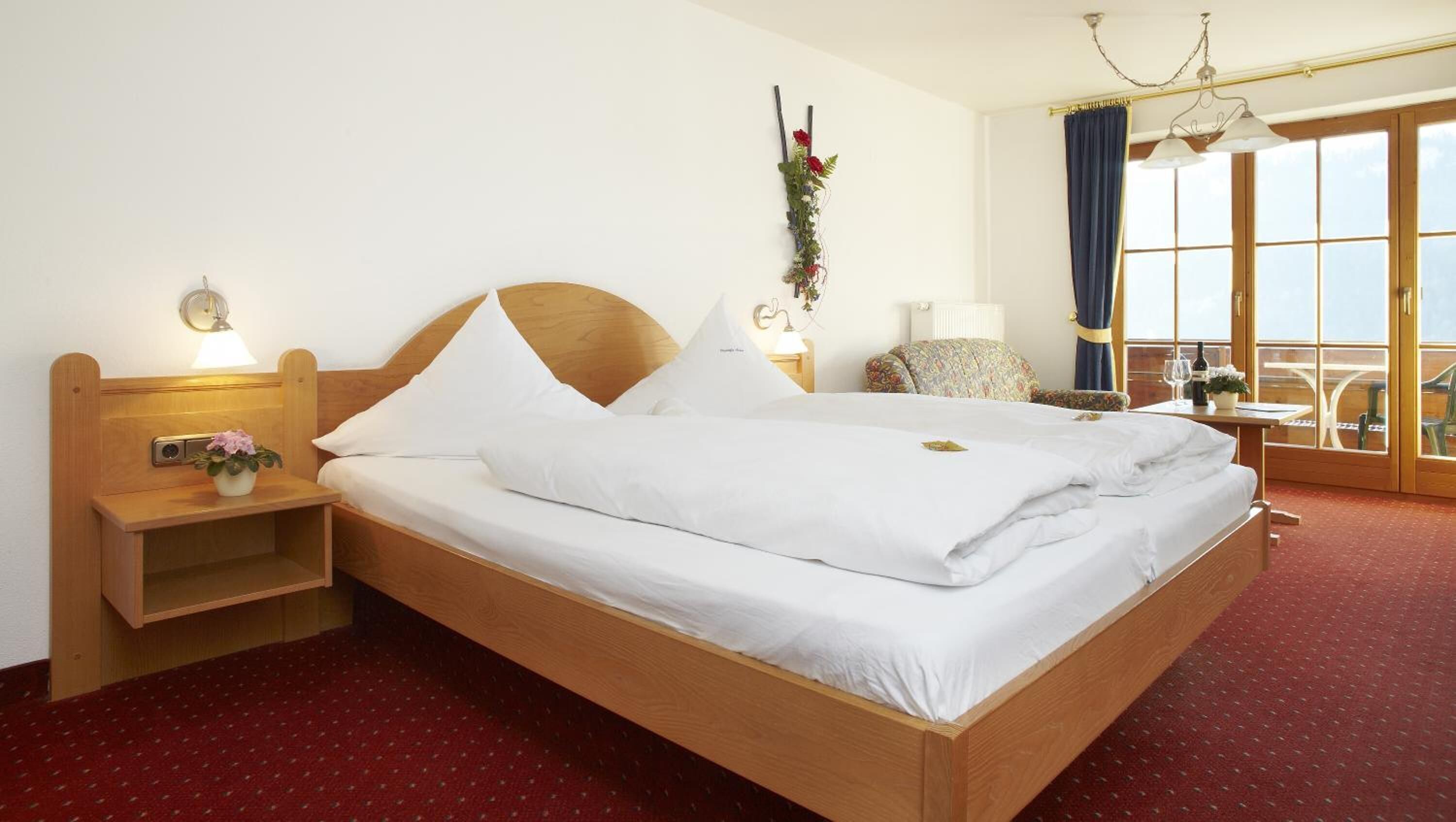 A double bed with white linens, a wooden headboard, a bedside table with a lamp, a chair, and a view of the outdoors through large windows.