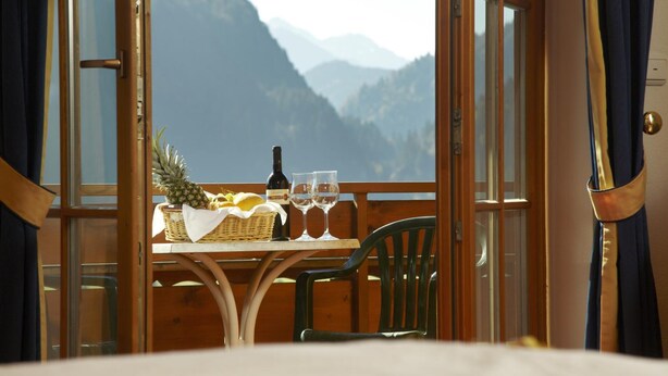 A table with a bottle, glasses, and a fruit basket, overlooking a mountainous landscape. - Hotel Oberdorfer Stuben (Obermaiselstein)