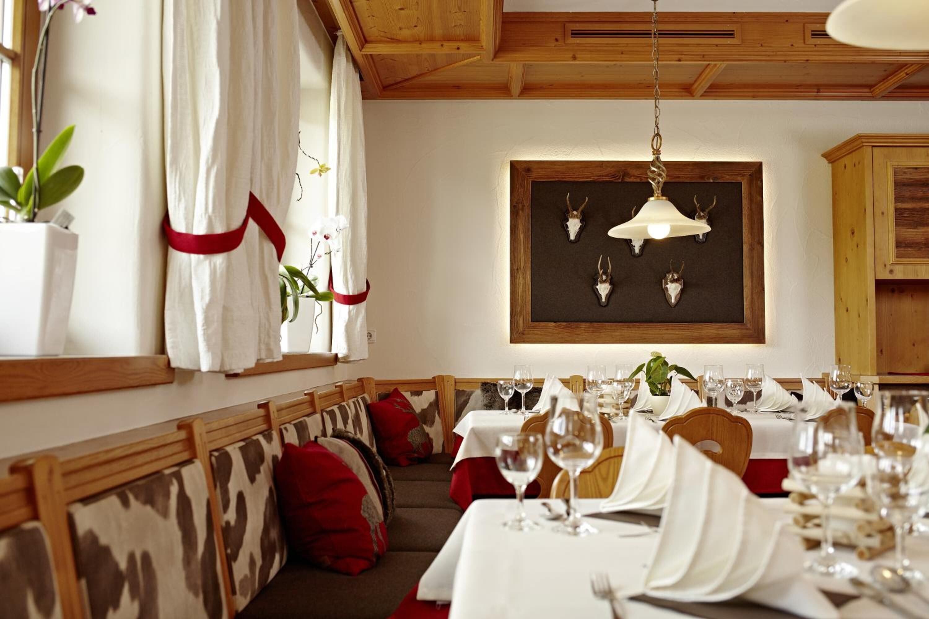 Comedor con bancos y mesas de madera dispuestas para una comida, con una araña y decoraciones de cráneos de animales en la pared.