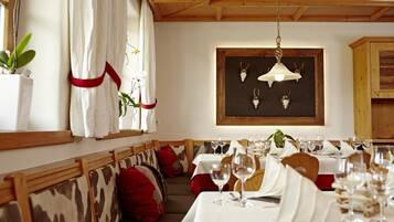 Comedor con bancos y mesas de madera dispuestas para una comida, con una araña y decoraciones de cráneos de animales en la pared.