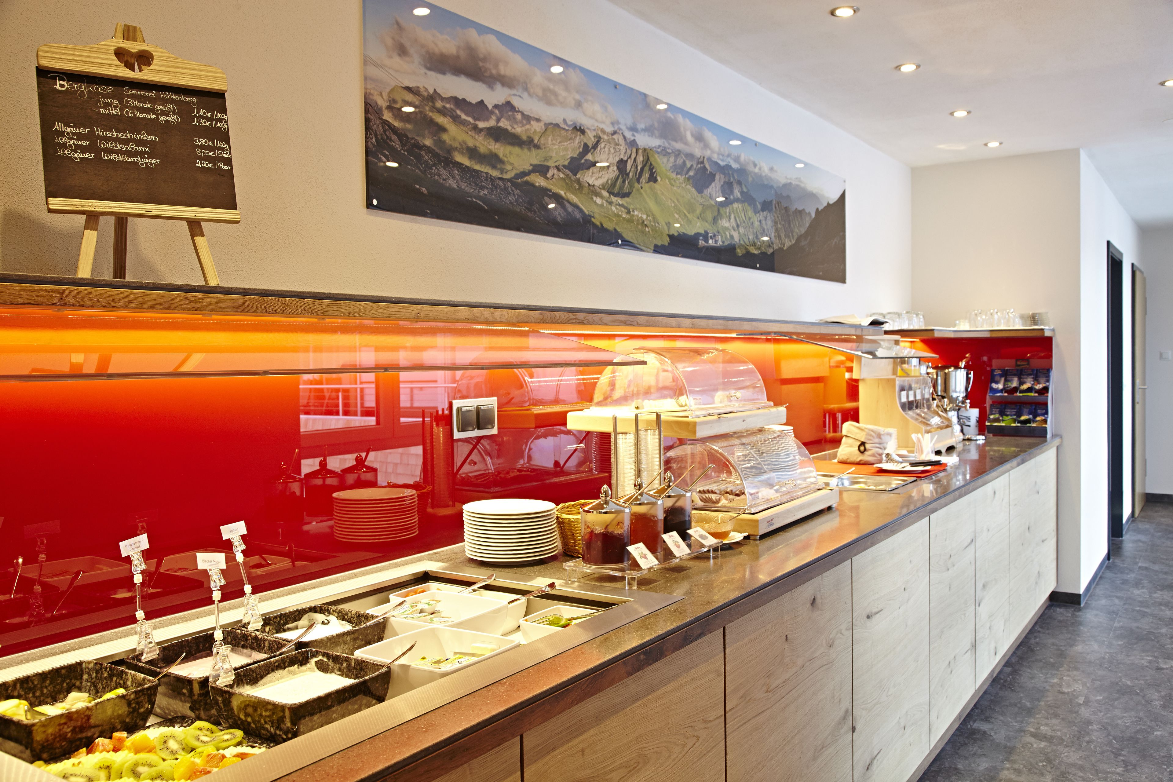 Un espace buffet proposant différents plats, un tableau affichant le menu et une peinture représentant un paysage de montagne au mur.