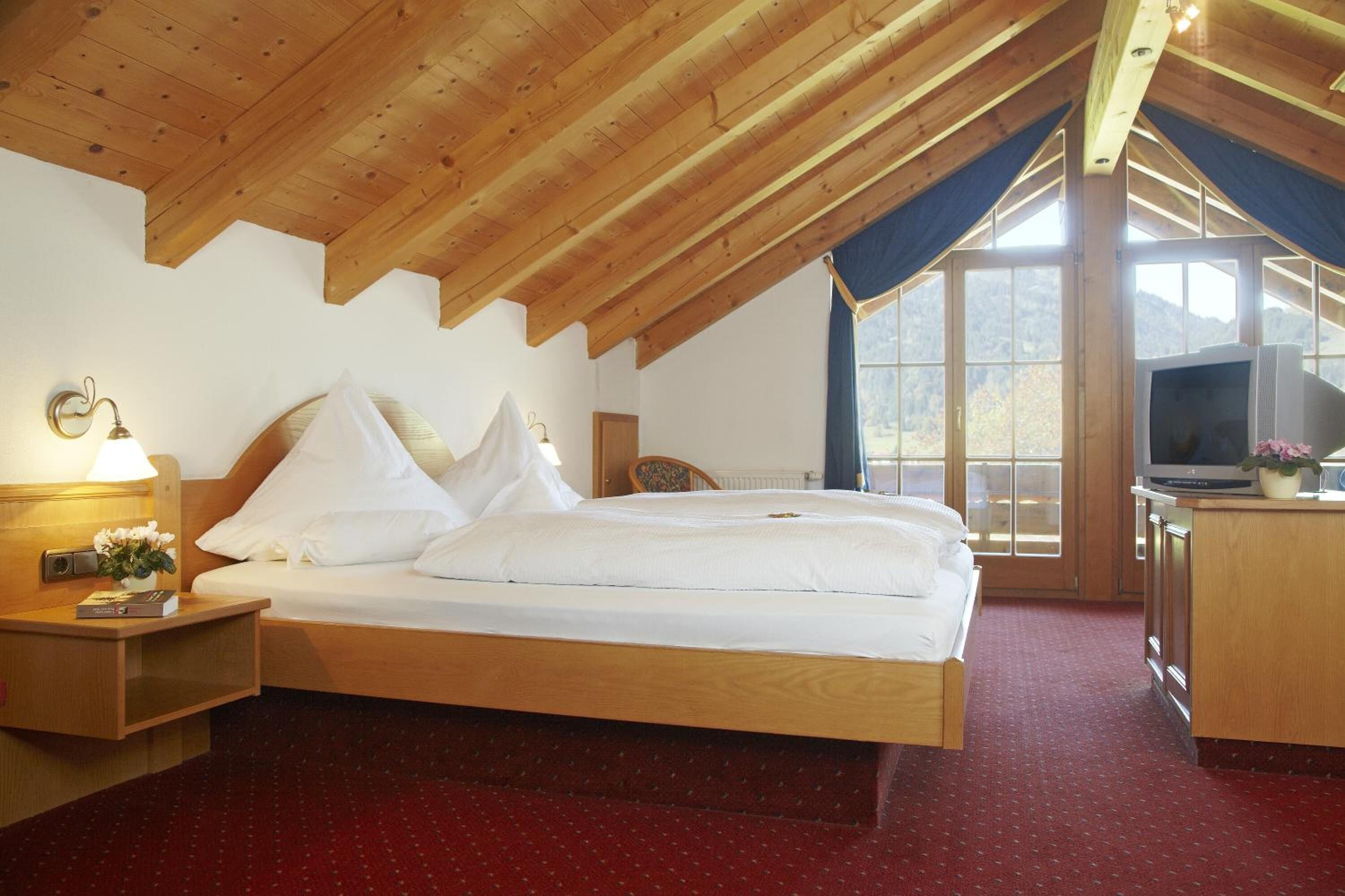 Una camera da letto con soffitto in legno, un letto con biancheria da letto bianca, un comodino con una lampada, una televisione e una finestra vista all'esterno.