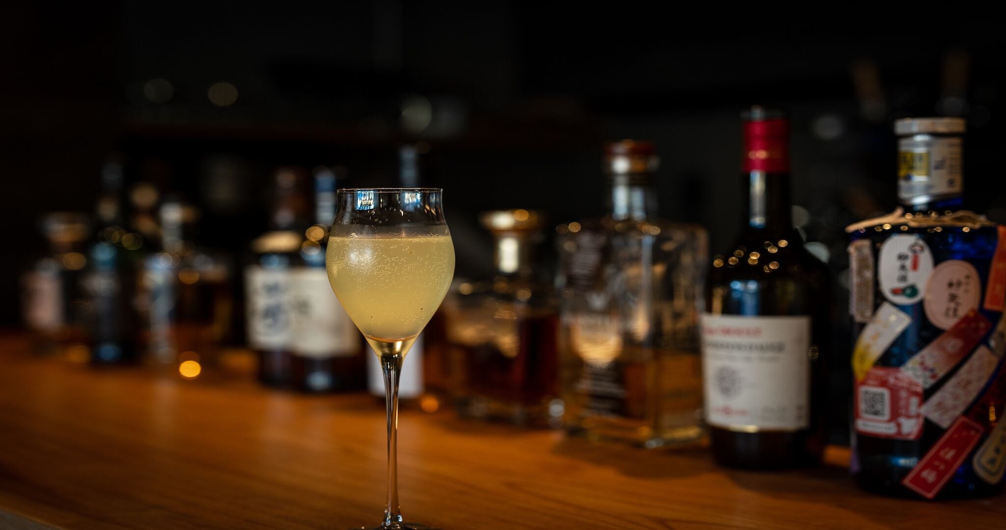 A bar with a glass of a light-colored beverage in the foreground, surrounded by various bottles and containers of alcohol.
