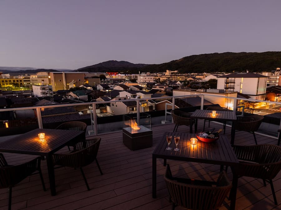 A rooftop terrace with outdoor seating, a fire feature, and a view of the cityscape at dusk.