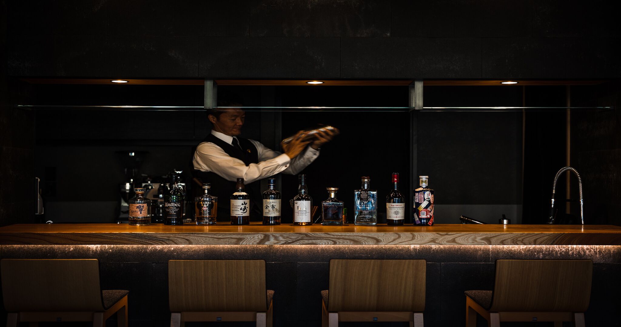 En bar med en bartender, der laver drinks, forskellige flasker med spiritus og barstole.