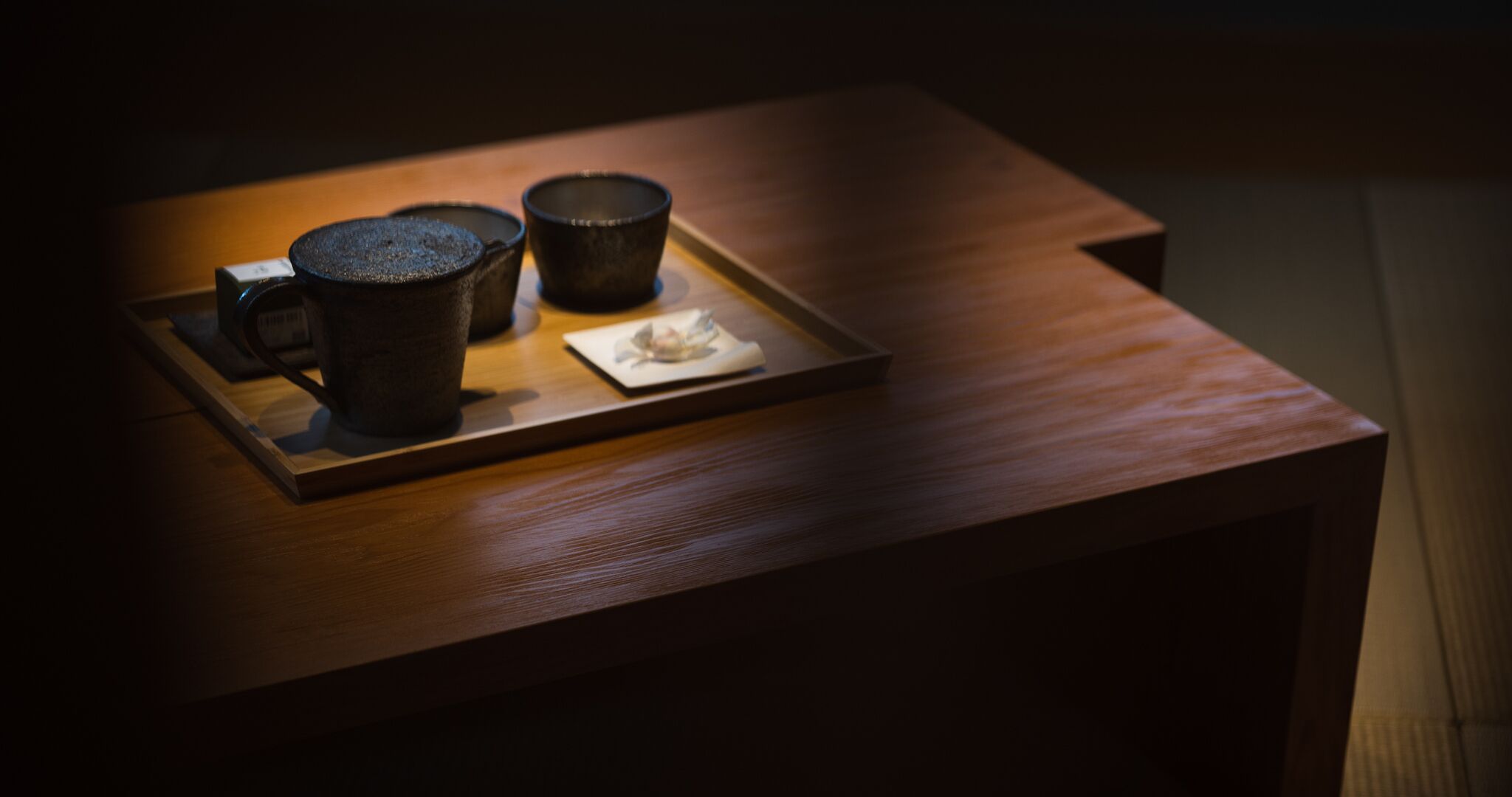A wooden table with a tray holding two dark ceramic cups and a small plate with a flower.