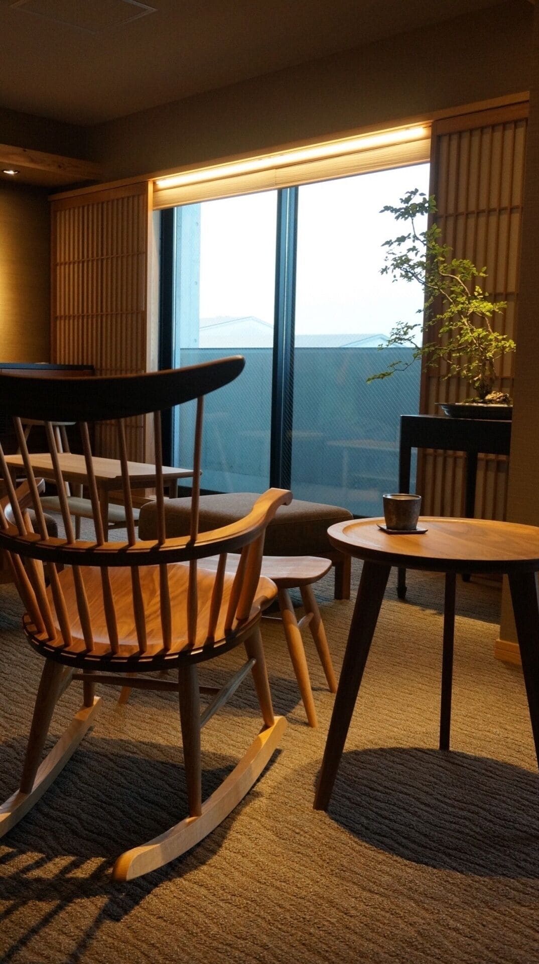 A modern hotel room with a large window offering a city view, a wooden rocking chair, a small table with a cup, and a potted plant.