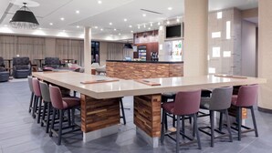 A modern bar area with a long wooden bar counter, multiple bar stools, and a seating area with chairs. - Courtyard by Marriott Cincinnati Mason (Mason)